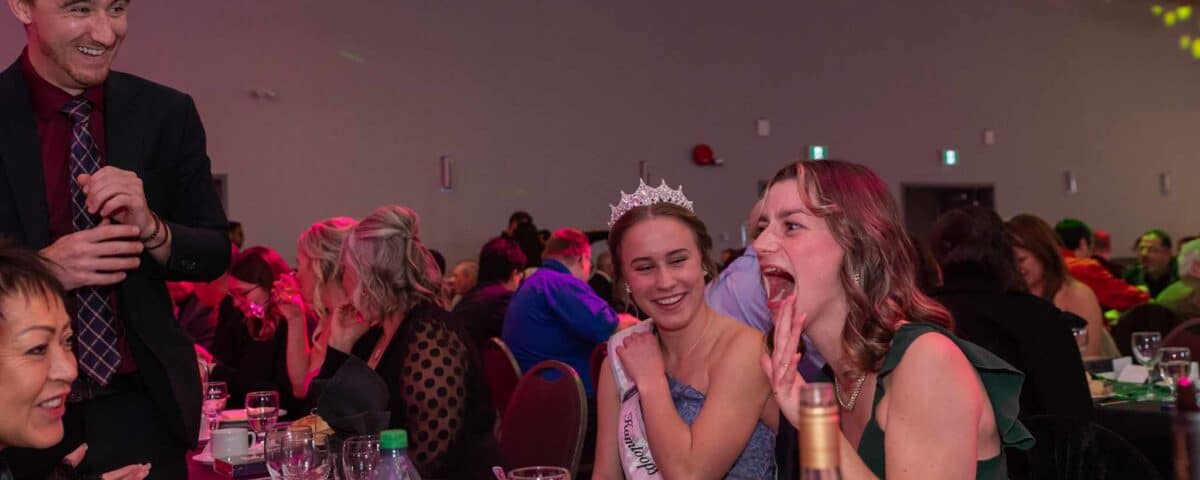 A festive gathering with guests at a dinner table, featuring a woman wearing a tiara and sash, laughing along with a friend, while a man engages with them.
