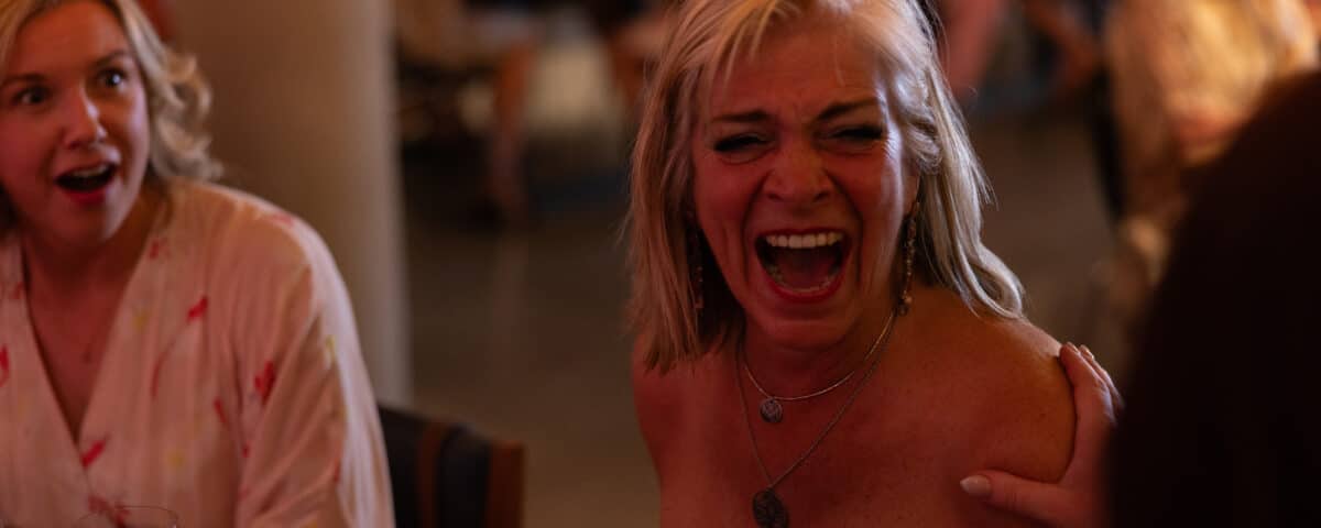 A woman with blonde hair is laughing joyfully at a social gathering, while another woman looks surprised. The background shows blurred figures and tables.