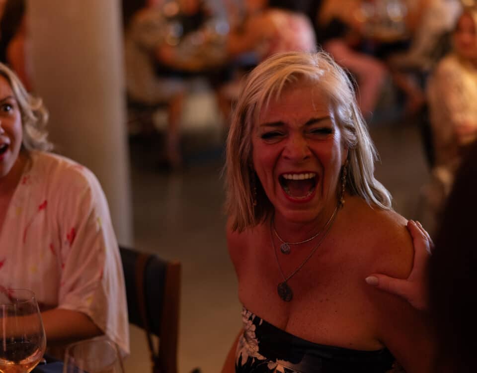A woman with blonde hair is laughing joyfully at a social gathering, while another woman looks surprised. The background shows blurred figures and tables.