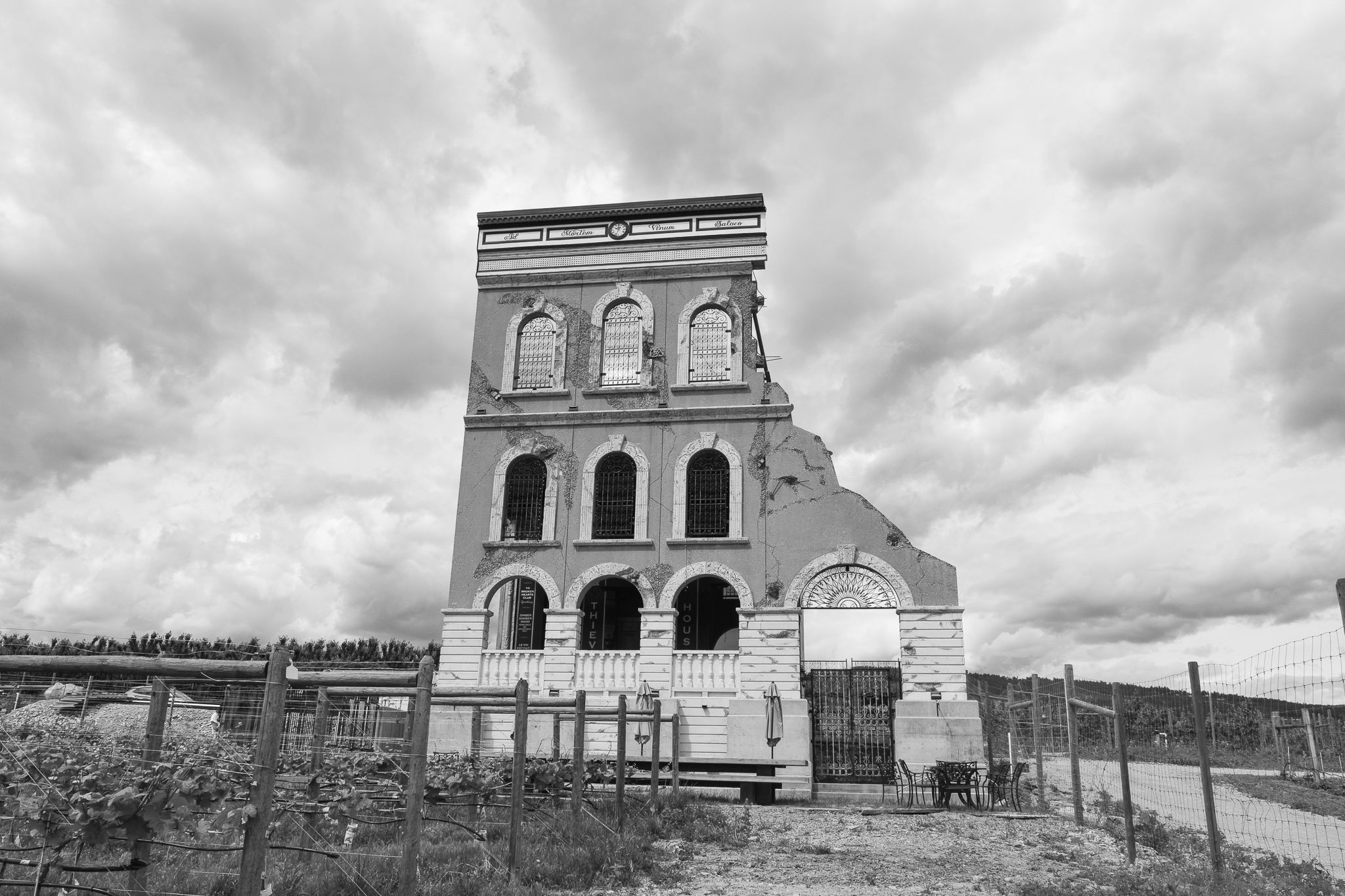 A partially collapsed, historical building with large arched windows, set against a cloudy sky, surrounded by a vineyard.