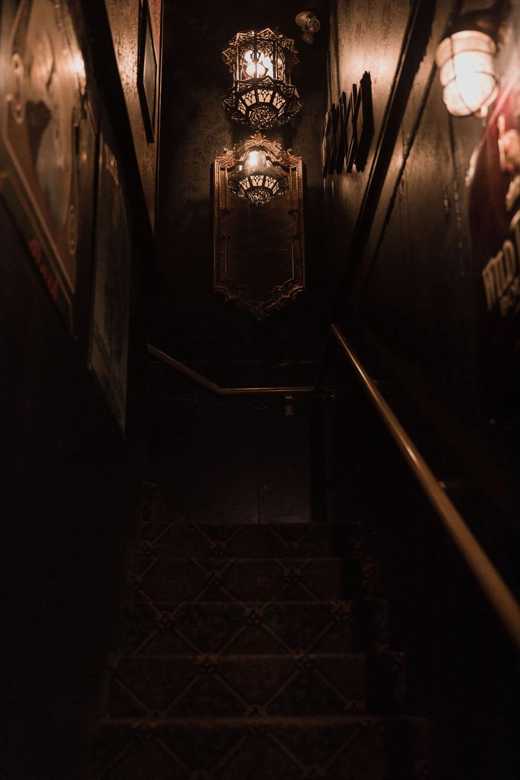 A dimly lit staircase leading downward, decorated with vintage-style wall lanterns and a mirror, surrounded by dark walls and framed artwork.