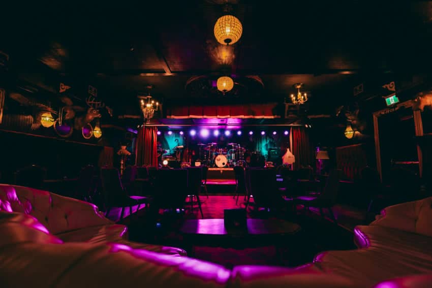 A dimly lit music venue with a stage, microphone, and instruments, surrounded by seating and decorative lighting.