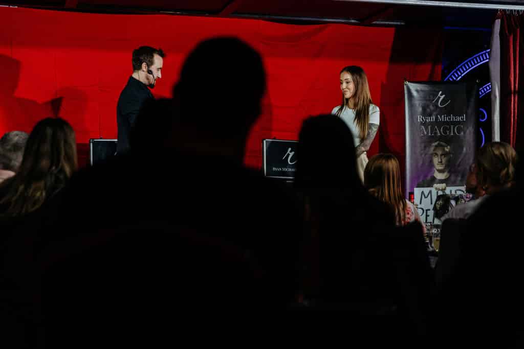 A magician performing on stage with an audience in silhouette; a woman stands beside him, and a promotional banner for Ryan Michael Magic is visible.
