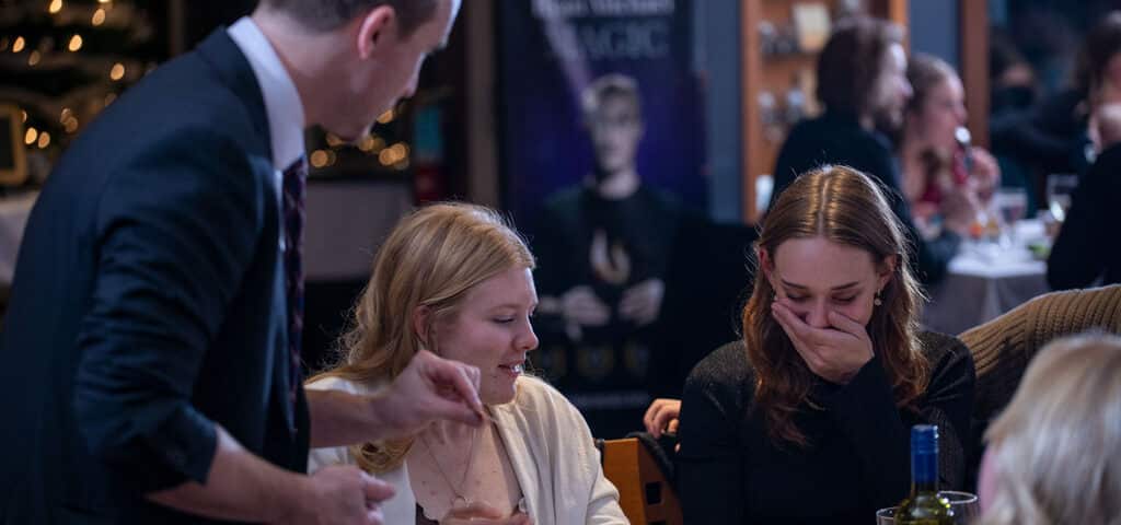 A magician performs at a dinner event, engaging with two women who are laughing and surprised. The setting includes tables with dinnerware and drinks, and a banner for a magic show in the background.