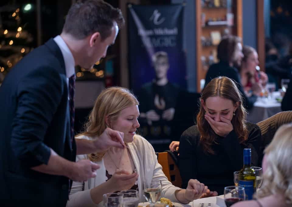 A magician performs at a dinner event, engaging with two women who are laughing and surprised. The setting includes tables with dinnerware and drinks, and a banner for a magic show in the background.