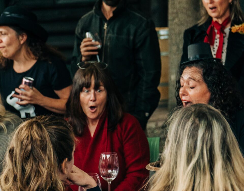 A lively gathering of people, some laughing and engaging, with a table featuring drinks and a festive atmosphere marked by colorful umbrellas overhead.