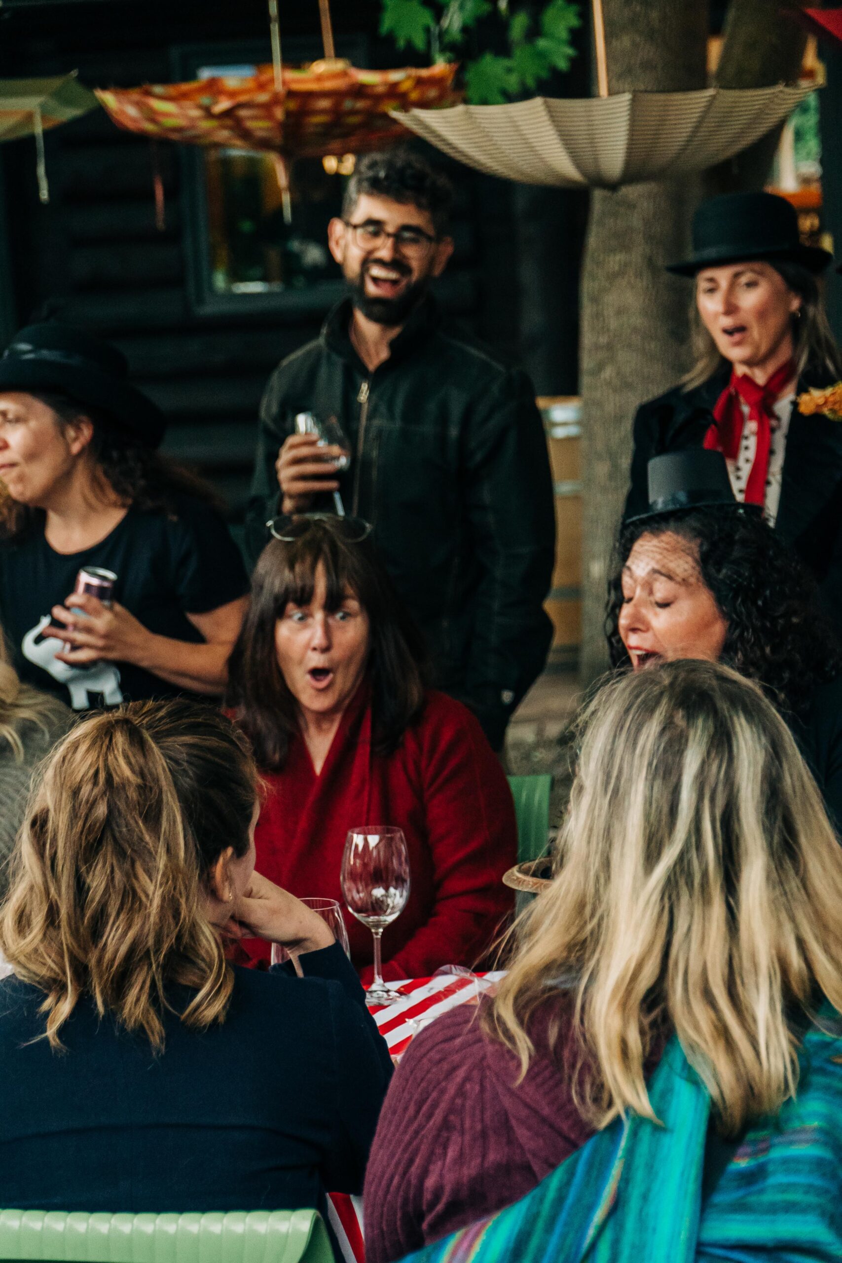A lively gathering of people, some laughing and engaging, with a table featuring drinks and a festive atmosphere marked by colorful umbrellas overhead.