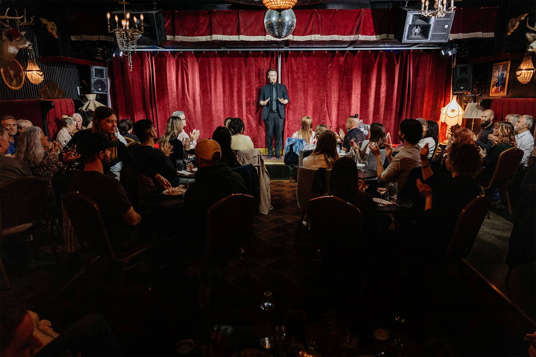 A performer stands on stage in a dimly lit venue with red curtains, while an audience, seated at tables, claps in appreciation.