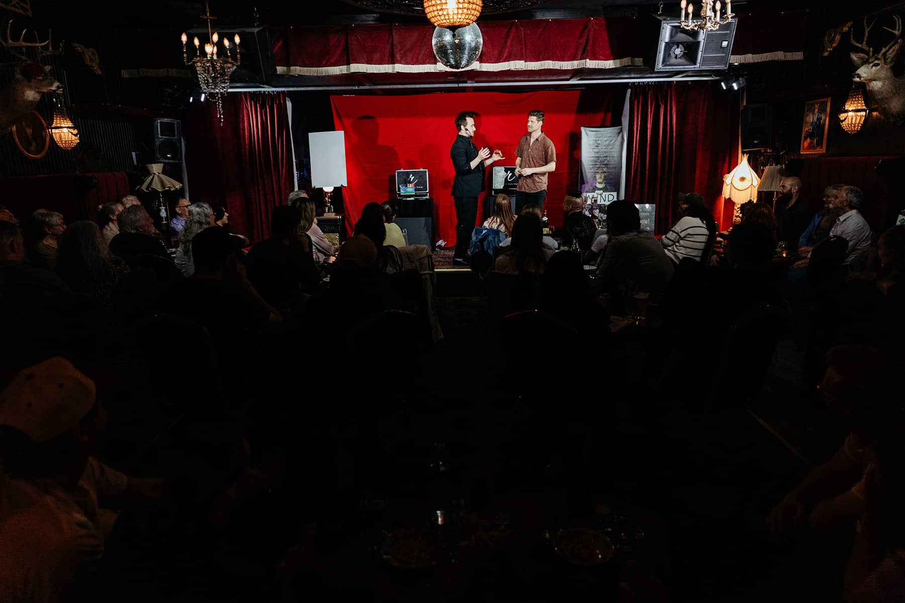 A stage performance with two presenters in front of an audience, featuring a red backdrop and various decorations, including chandeliers and framed images.