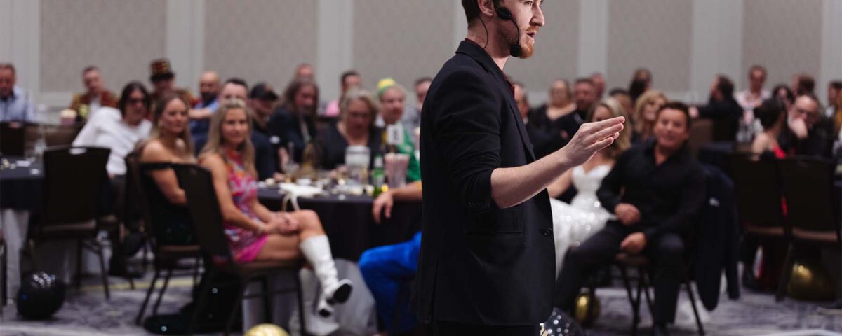 A speaker with a microphone gestures while addressing an audience at a lively event, with tables and decorations in the background.