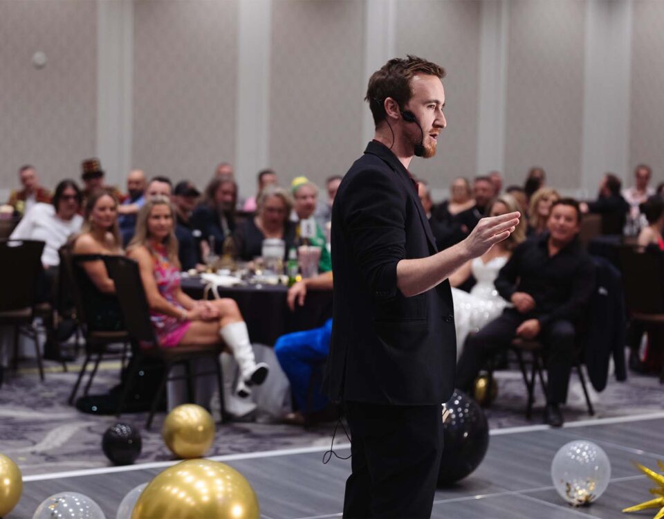 A speaker with a microphone gestures while addressing an audience at a lively event, with tables and decorations in the background.