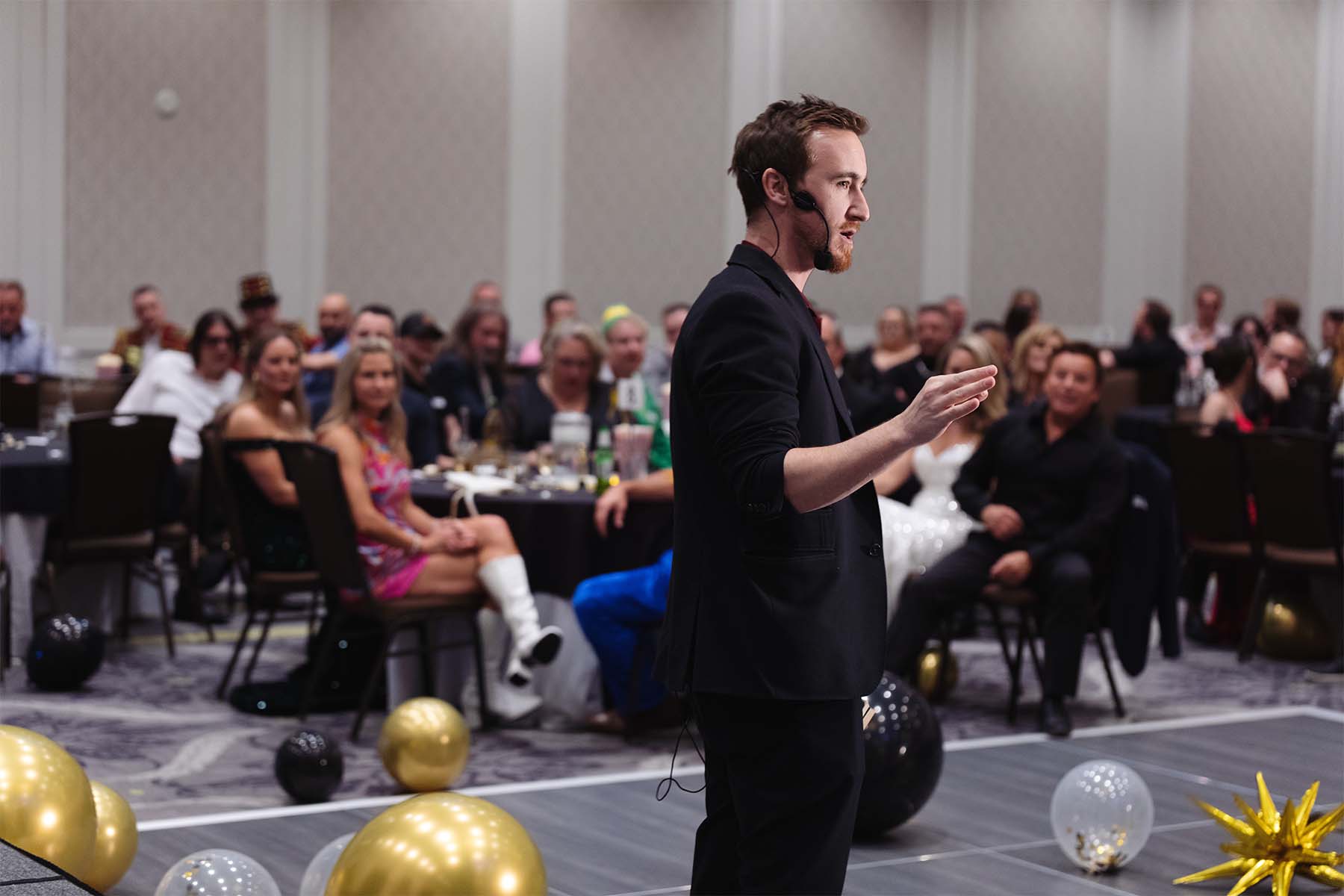 A speaker with a microphone gestures while addressing an audience at a lively event, with tables and decorations in the background.