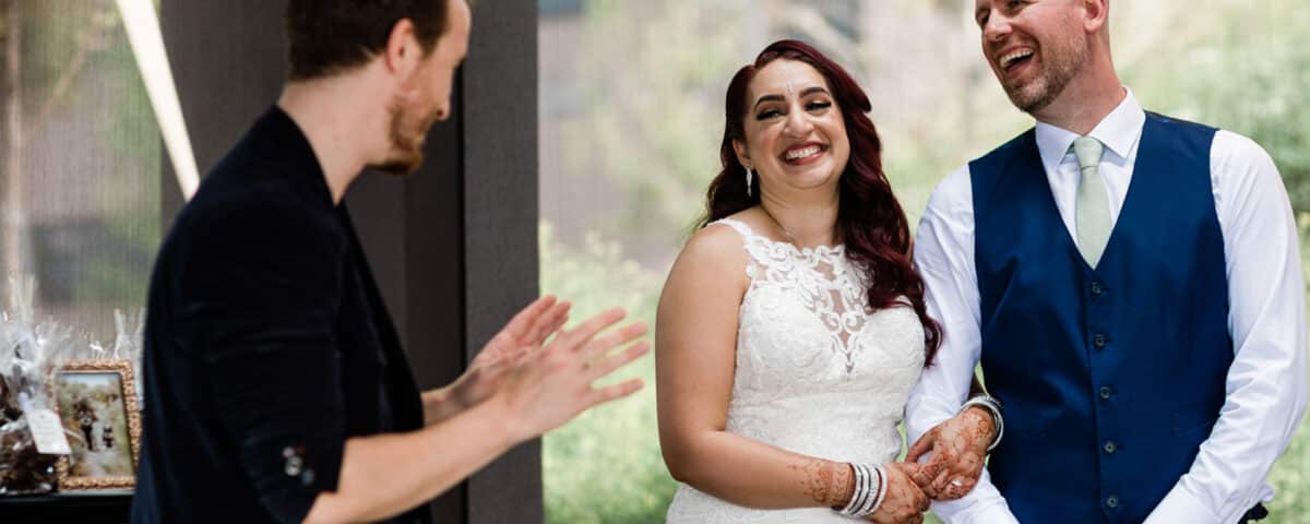 A bride and groom smile at each other during a toast or speech, with a person speaking in front of them. The setting appears elegant, with greenery visible in the background.