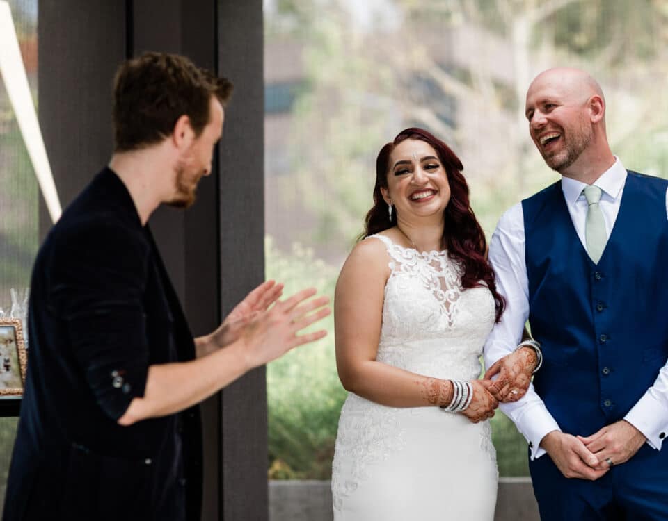 A bride and groom smile at each other during a toast or speech, with a person speaking in front of them. The setting appears elegant, with greenery visible in the background.