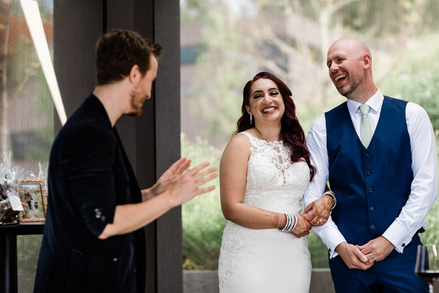 A bride and groom smile at each other during a toast or speech, with a person speaking in front of them. The setting appears elegant, with greenery visible in the background.