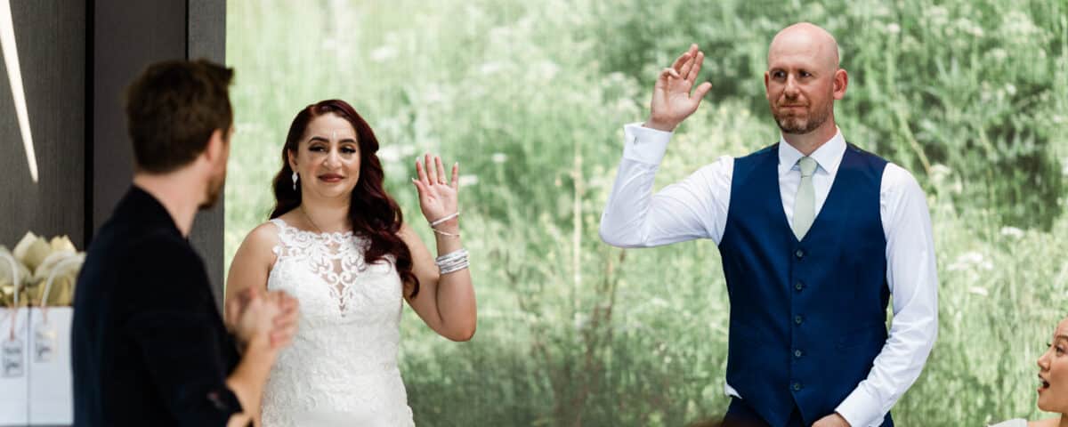 A bride and groom raising their hands during a celebration, with guests seated at a table nearby. The background features greenery.