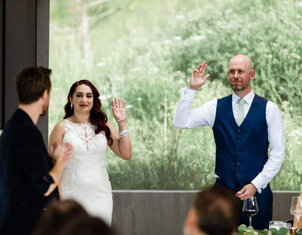 A bride and groom raising their hands during a celebration, with guests seated at a table nearby. The background features greenery.