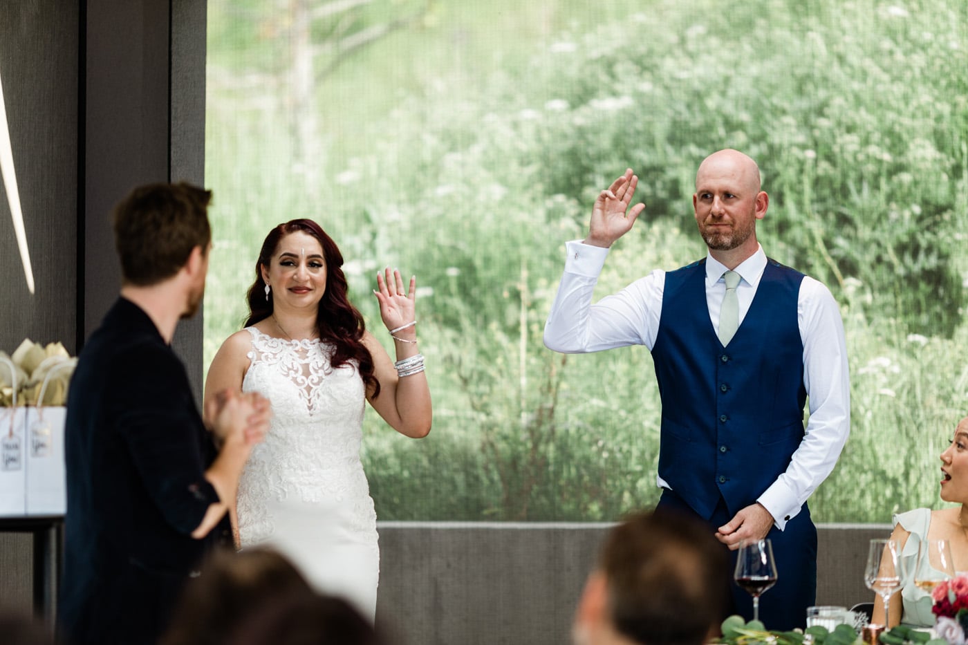 A bride and groom raising their hands during a celebration, with guests seated at a table nearby. The background features greenery.
