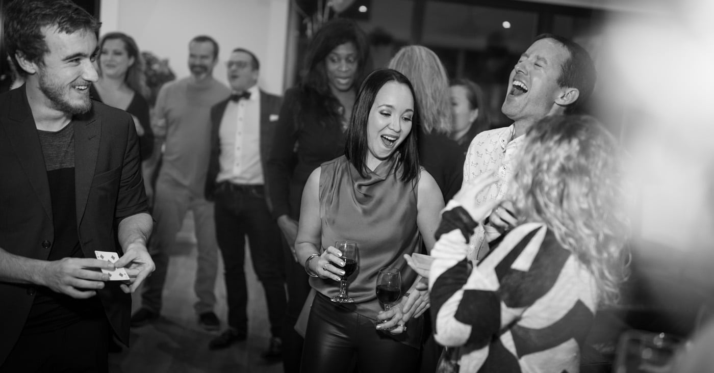 A lively social gathering with people laughing and enjoying themselves, some holding drinks. One person is performing a magic trick, and others are watching and interacting.