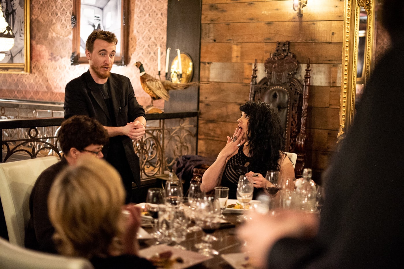 A man in a black jacket is speaking animatedly to a table of guests in a dimly lit restaurant, while a woman with curly dark hair reacts with surprise