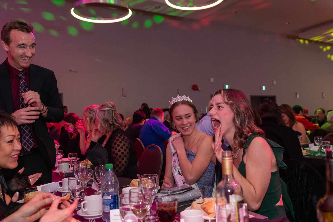 A lively social event at a banquet table with guests enjoying food and drinks; a smiling man in a suit interacts with two women, one wearing a tiara and another laughing. Colorful lights create a festive atmosphere.