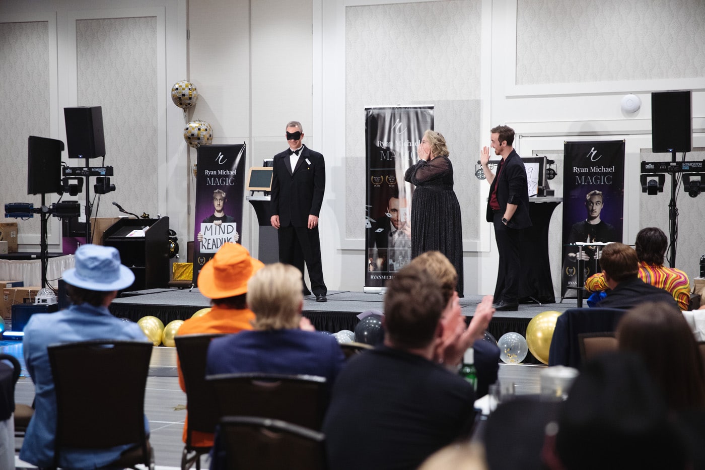 A magician performing on stage, accompanied by two assistants and an audience in formal attire. Banners promoting Ryan Michael's magic show are displayed in the background.