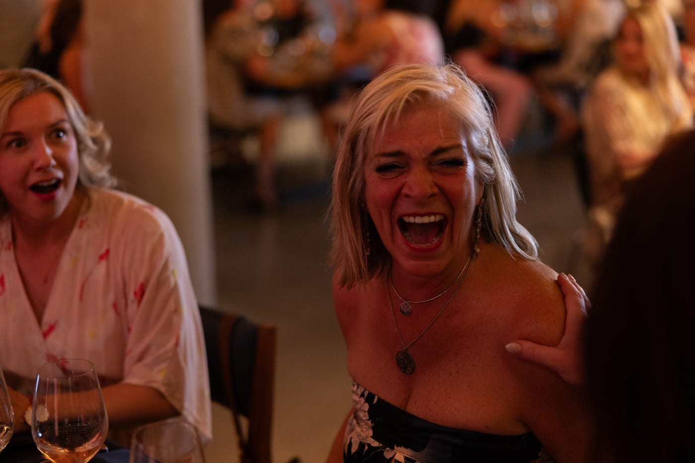 A woman with long, blonde hair is laughing joyfully while sitting at a table. Another woman, looking surprised, is seated nearby.