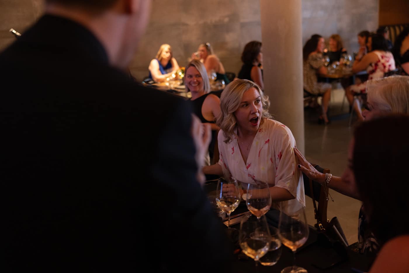A woman with a surprised expression sits at a table surrounded by others, with glasses of wine. The setting appears to be a social event or gathering.