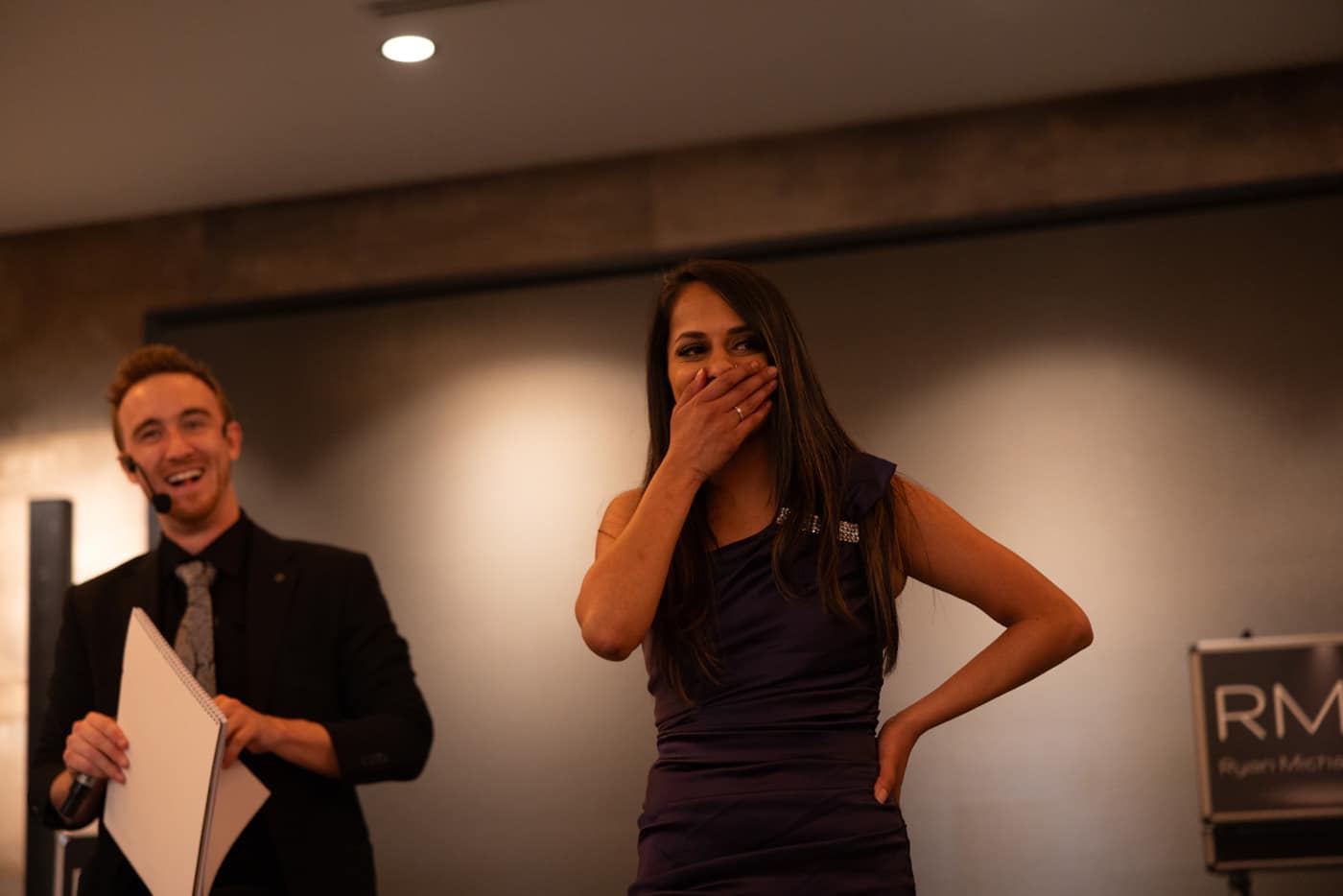 A man in a black suit with a microphone stands next to a woman in a purple dress, who is laughing and covering her mouth, during an event.