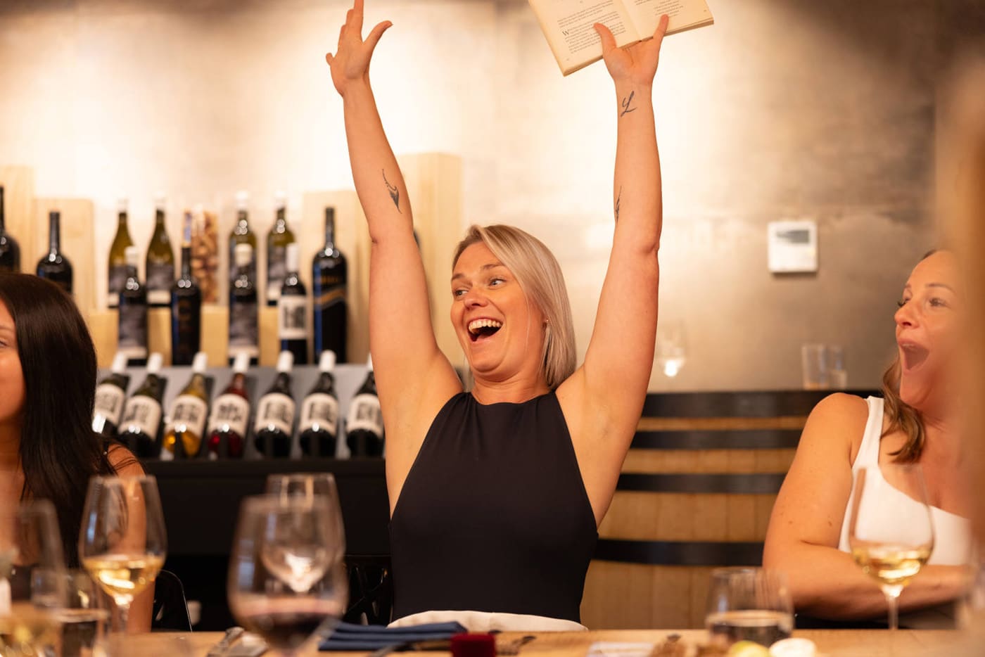 A woman with blonde hair is smiling and raising her arms excitedly in a restaurant setting, with shelves of wine bottles in the background.