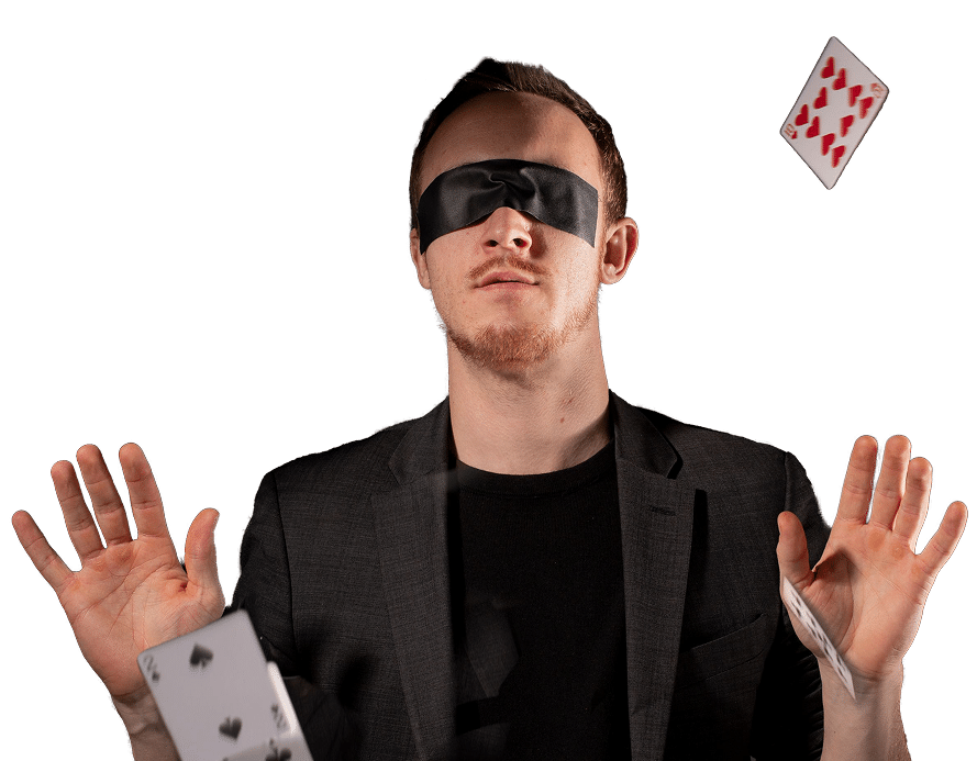 A man wearing a blindfold, dressed in a black jacket, is surrounded by flying playing cards, with his hands raised as if in a magical or theatrical gesture.