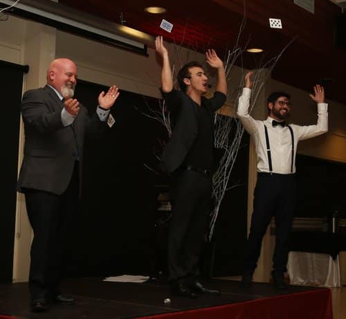 Three male performers are on stage, with arms raised, performing a magic trick. They appear to be engaged with an audience.