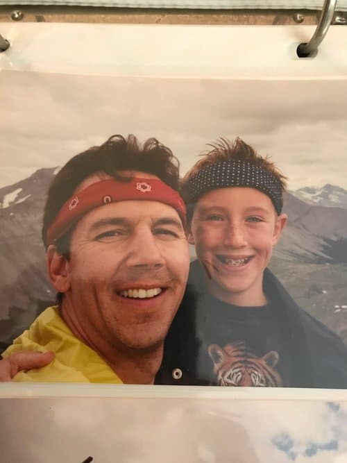 A man and a boy are smiling together outdoors, with mountains in the background.