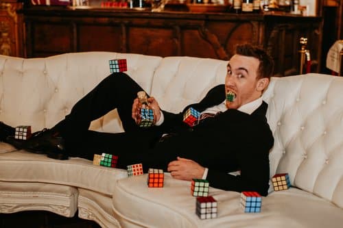 A man in a suit is lounging on a white couch, holding a Rubik's Cube in his mouth, surrounded by several other Rubik's Cubes scattered around him.
