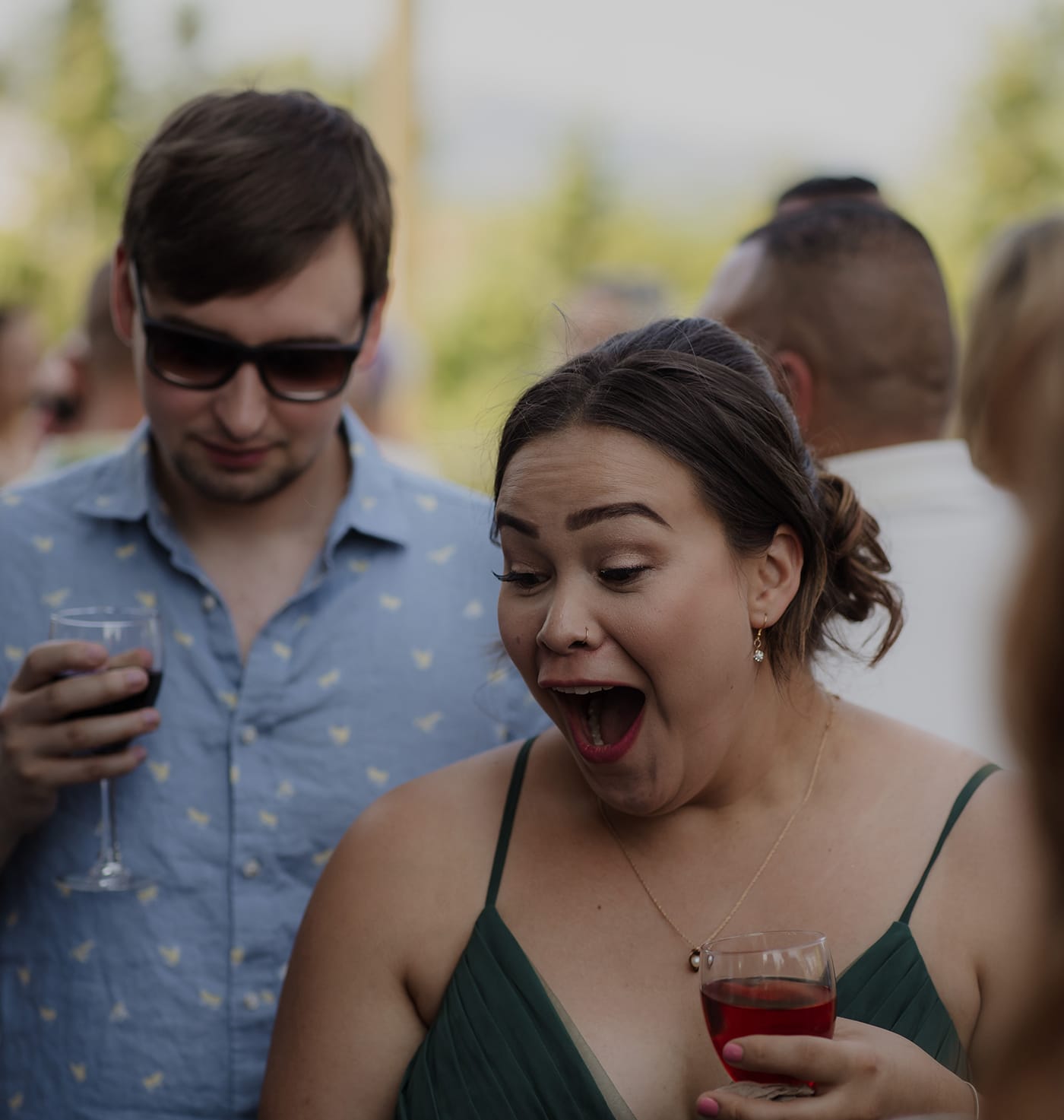 A woman in a green dress expresses surprise while holding a drink, with a man in a blue shirt and sunglasses looking on.