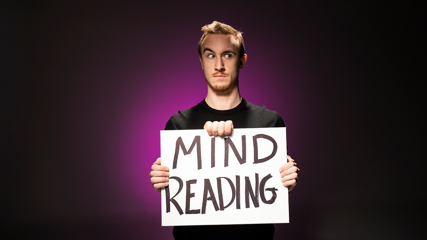 A person holding a sign that says "MIND READING," against a dark background with a purple light.