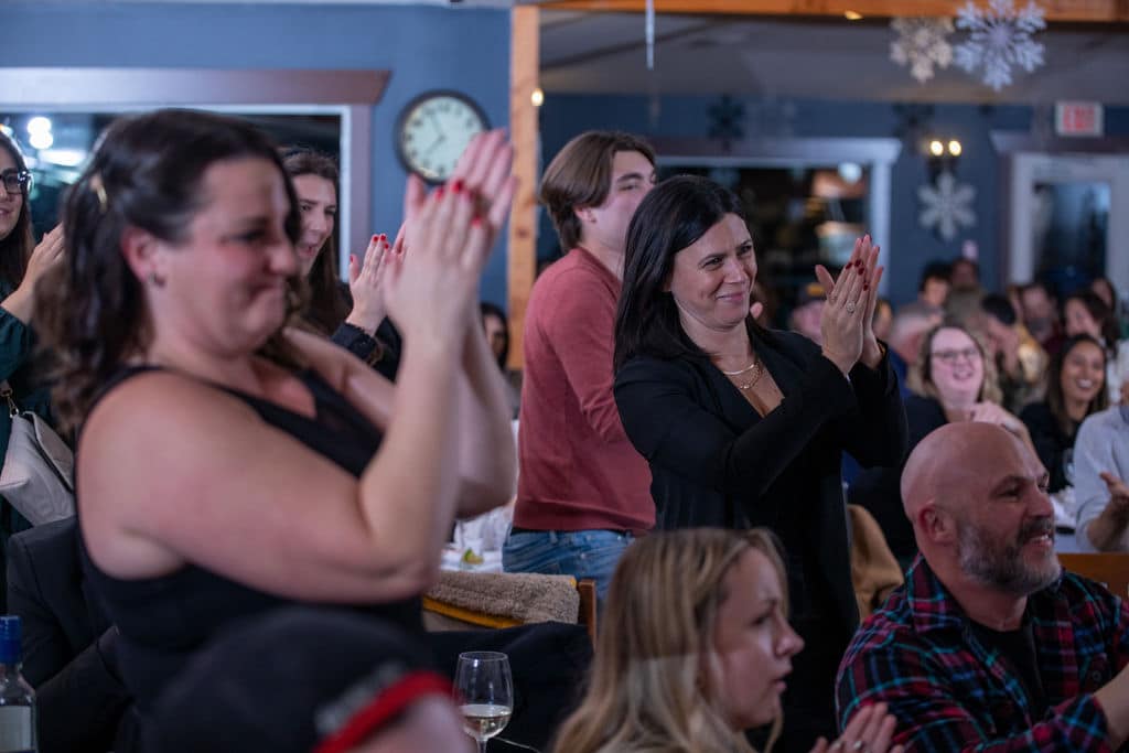 A crowd of people applauding in a venue. Some individuals are smiling and engaged, while others are clapping their hands.