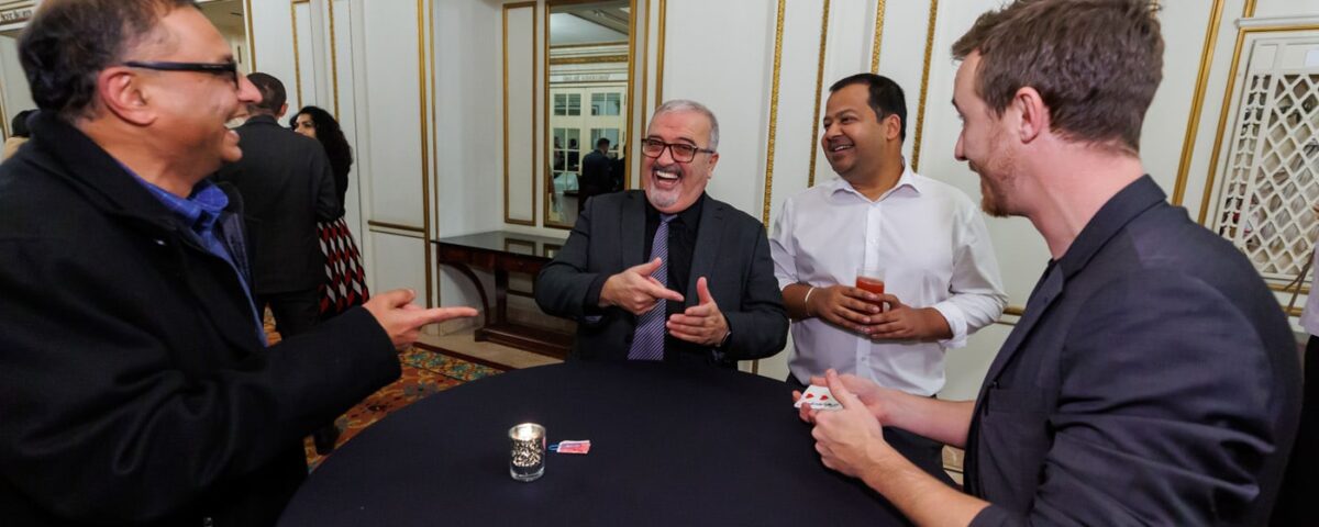 A group of four men are engaging in a lively conversation around a table, with a deck of cards and drinks in front of them, in an elegant room with decorative details.