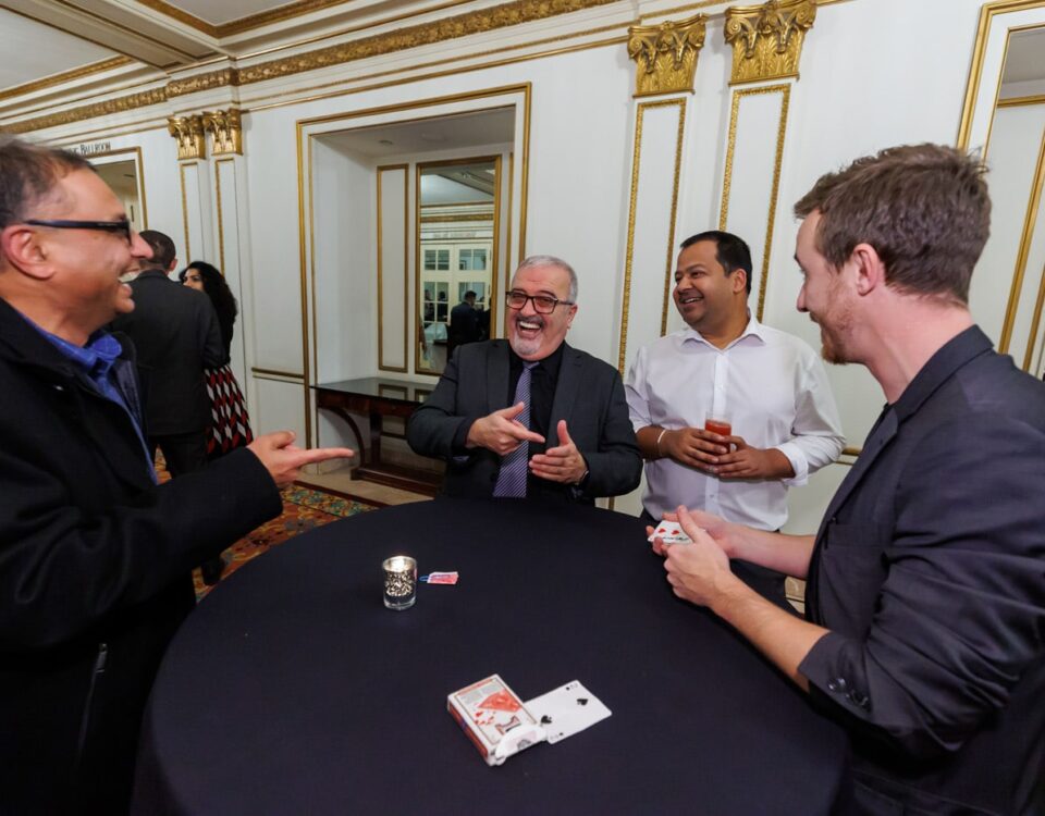 A group of four men are engaging in a lively conversation around a table, with a deck of cards and drinks in front of them, in an elegant room with decorative details.