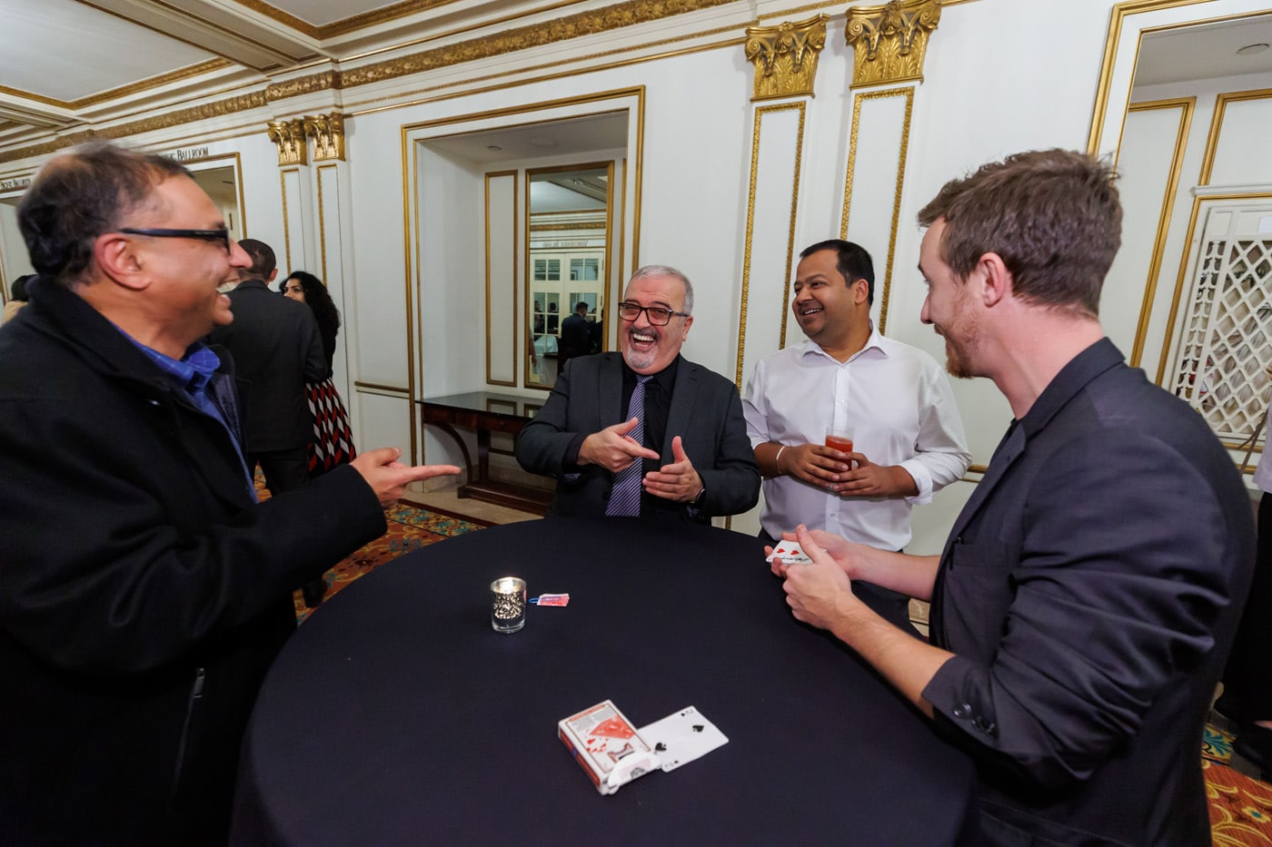 A group of four men are engaging in a lively conversation around a table, with a deck of cards and drinks in front of them, in an elegant room with decorative details.