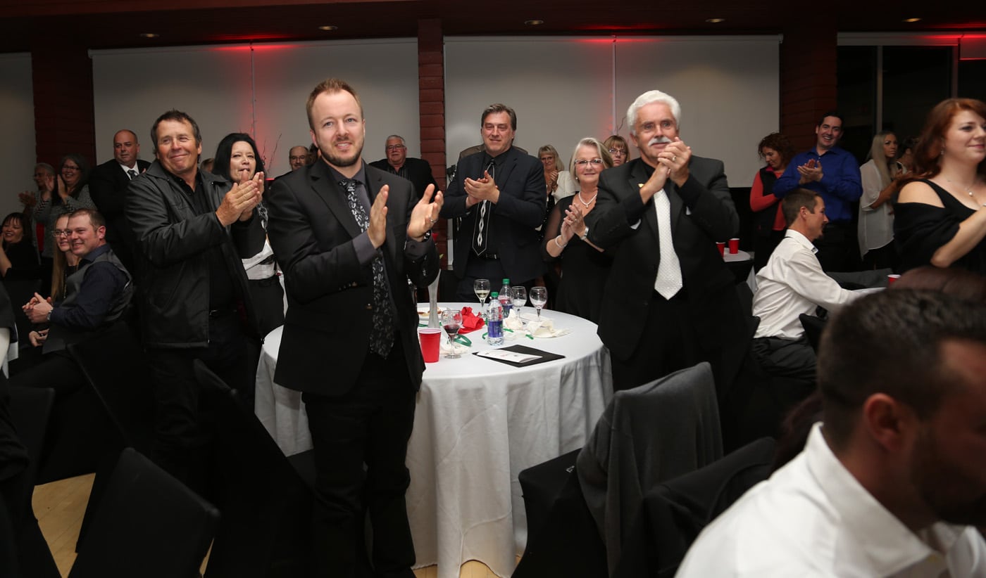 A group of people is clapping and standing, celebrating at an event. Tables are set with drinks and plates in a well-lit venue.