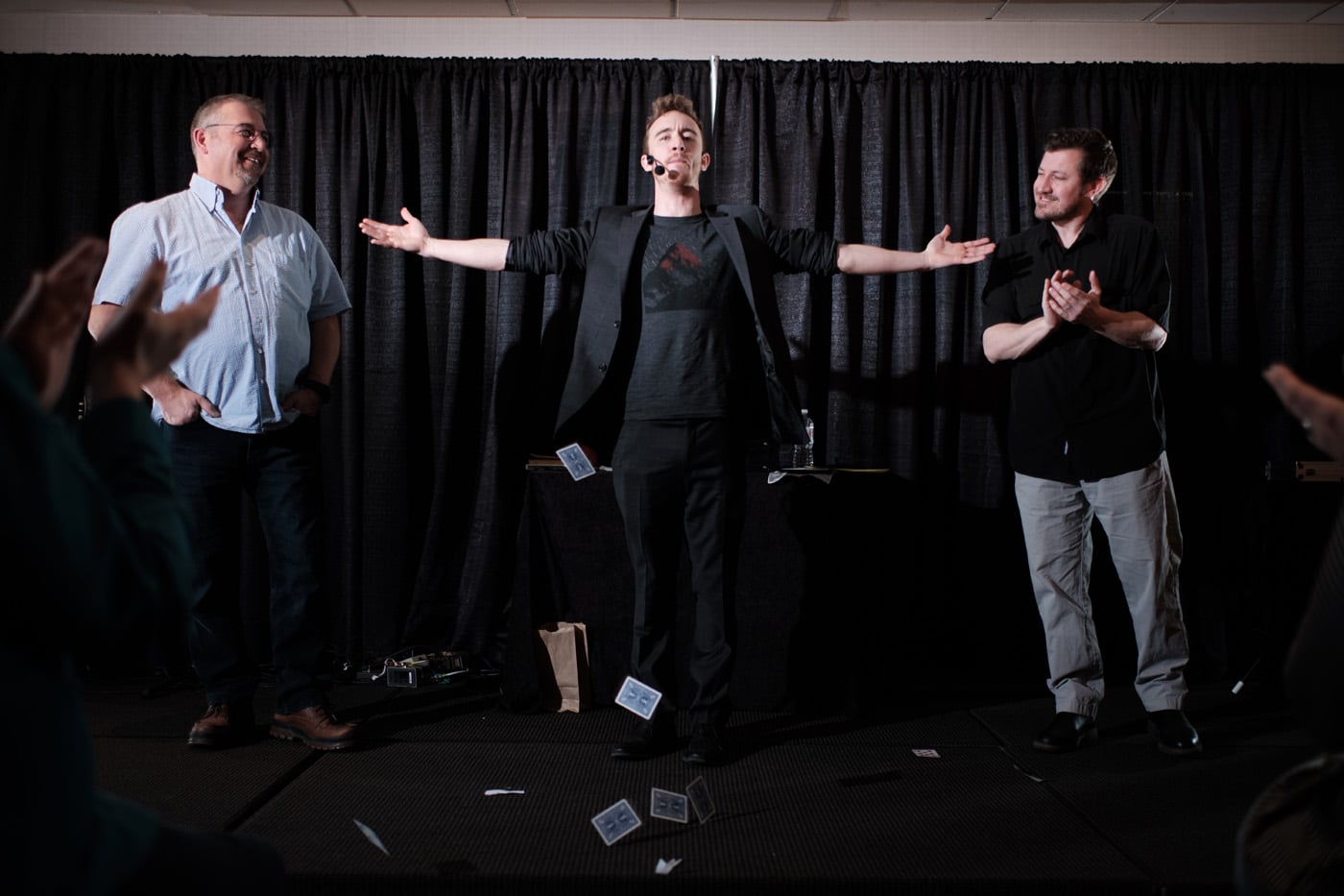 A magician performs on stage with two assistants, while the audience applauds. Cards are falling around the magician.