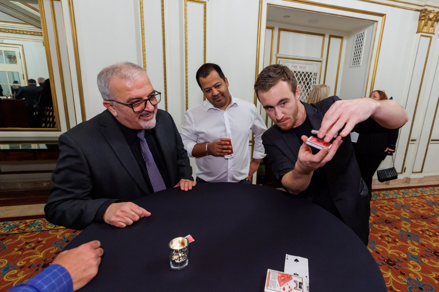 Four people are gathered around a table, watching a magician perform a card trick. One man in a black suit is smiling, while another, wearing a white shirt, holds a drink.