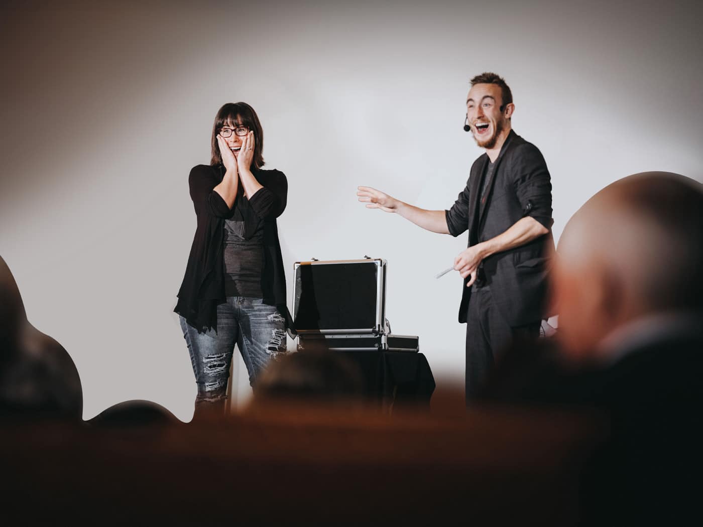 A man in a black suit is performing on stage, smiling and gesturing, while a woman in casual attire looks surprised and excited, standing beside him. A black case is visible on the stage.