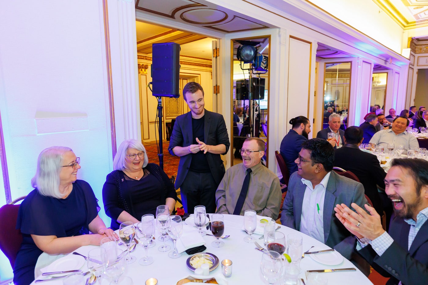 A round table at a formal event with several attendees enjoying a magician's performance. Laughter and conversation are taking place among the guests.