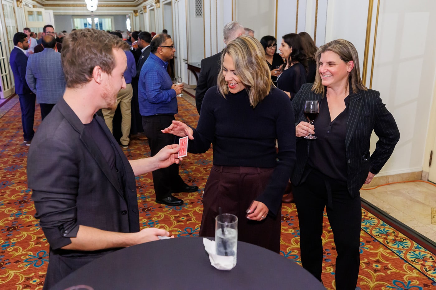 A group of people socializing at an event. A man performs a card trick for two women, one of whom is laughing and receiving a card, while another woman holds a glass of wine.