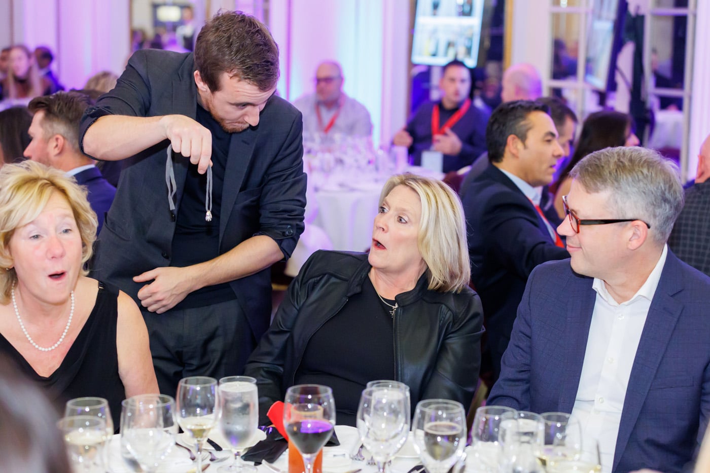 A magician performs at a dinner event, surprising a woman in a black jacket while a man in a suit looks on. Glasses of drinks are on the tables.