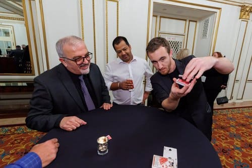 A magician performing a card trick for an audience at a table, with engaged spectators watching.