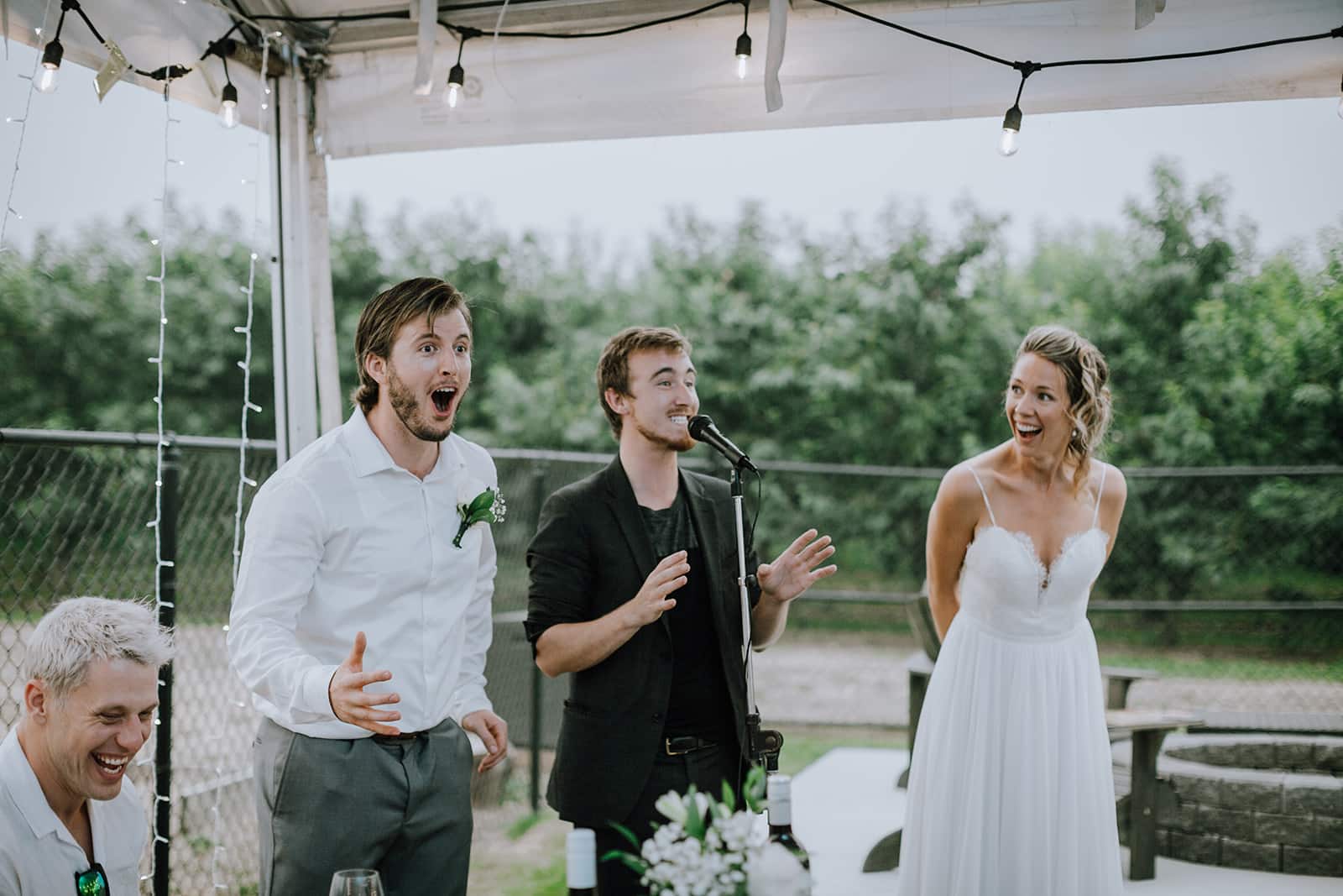 A joyful group of people at a wedding celebration, featuring a man with an open-mouthed expression, a singer at a microphone, and a smiling bride. The setting has string lights and an outdoor ambiance.