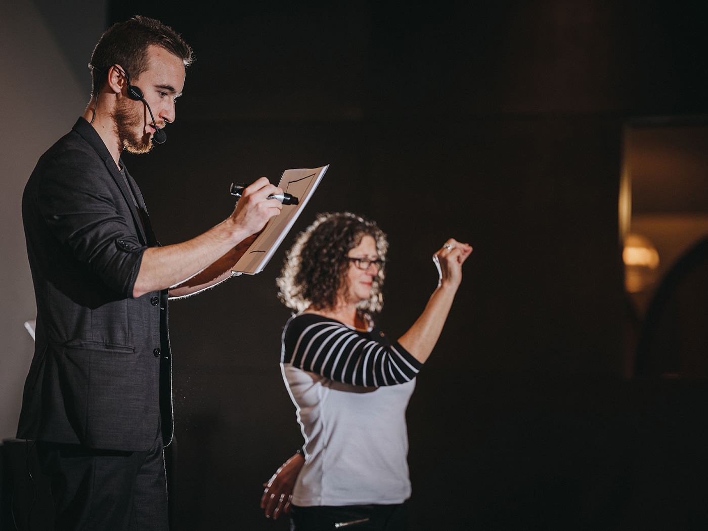 A man with a microphone is drawing on paper, while a woman in a striped shirt is gesturing.